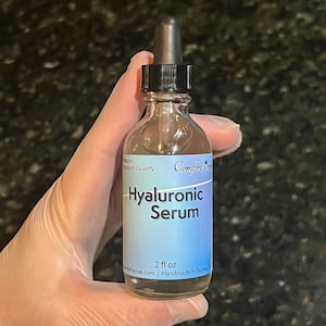 May include: A clear glass bottle of Hyaluronic Serum with a black dropper cap. The light blue label features the text Comfort Sense, Hyaluronic Serum, and 2 fl oz. The bottle contains a transparent liquid and is held upright against a dark background.