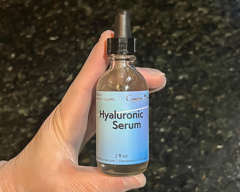 May include: A clear glass bottle of Hyaluronic Serum with a black dropper cap. The light blue label features the text Comfort Sense, Hyaluronic Serum, and 2 fl oz. The bottle contains a transparent liquid and is held upright against a dark background.