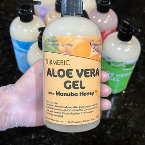 May include: A clear bottle of turmeric aloe vera gel with Manuka honey, held in a gloved hand. The label reads "All Natural Premium Quality" and "Comfort Sense." Other bottles of gel are visible in the background. The bottle contains 16 fl oz.