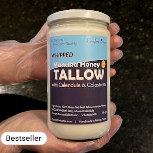May include: A white jar with a black label that reads "Whipped Manuka Honey Tallow with Calendula & Colostrum". The jar is labeled "All Natural Premium Quality" and "Comfort Sense". The jar is 24 oz and contains milk. The jar is being held by a gloved hand. The label also says "Handmade in Palmer, Texas".