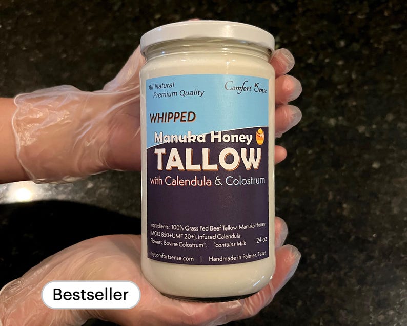 May include: A white jar with a black label that reads "Whipped Manuka Honey Tallow with Calendula & Colostrum". The jar is labeled "All Natural Premium Quality" and "Comfort Sense". The jar is 24 oz and contains milk. The jar is being held by a gloved hand. The label also says "Handmade in Palmer, Texas".