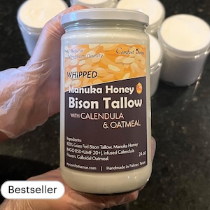 May include: A glass jar of whipped Manuka honey and bison tallow body butter with calendula and oatmeal. The jar is labeled "Whipped Manuka Honey Bison Tallow with Calendula & Oatmeal". The label also includes the text "All Natural Premium Quality" and "Comfort Sense".