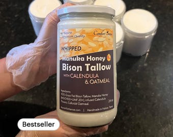 Puede incluir: Un tarro de vidrio de mantequilla corporal batida de miel de Manuka y sebo de bisonte con caléndula y avena. El tarro está etiquetado como "Whipped Manuka Honey Bison Tallow with Calendula & Oatmeal". La etiqueta también incluye el texto "All Natural Premium Quality" y "Comfort Sense".