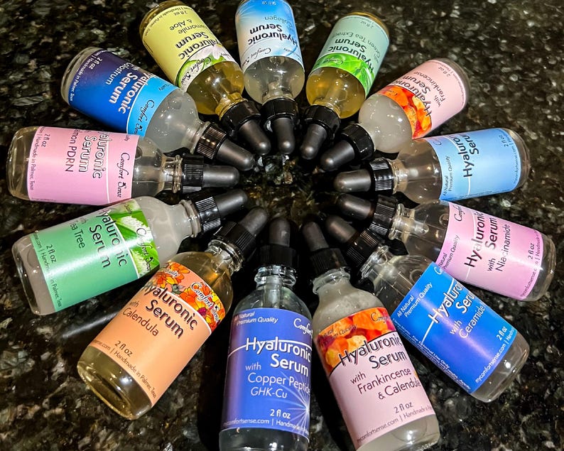 May include: A circular arrangement of twelve clear glass dropper bottles containing Hyaluronic Serum. Each bottle features a colored label with specific ingredient names like Calendula, Niacinamide, and Copper Peptide, all marked as 2 fl oz.