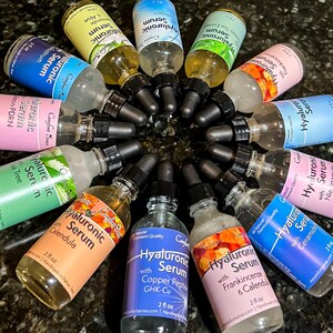 May include: A collection of twelve glass dropper bottles containing Hyaluronic Serum in various colors, each labeled with 2 fl oz volume and specific ingredients like Chamomile, Green Tea, Frankincense, Niacinamide, Ceramide, and Copper Peptide.