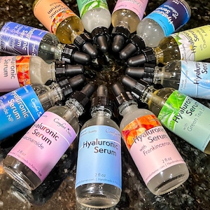 May include: A circular arrangement of twelve clear glass dropper bottles containing Hyaluronic Serum. Each bottle features a black dropper cap and a label with specific ingredients like Niacinamide, Frankincense, Collagen, and Calendula, each holding 2 fl oz.