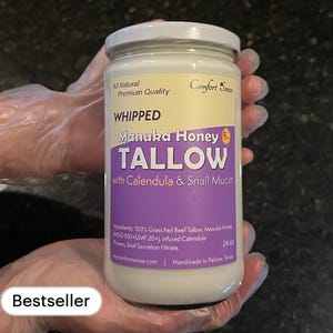 May include: A glass jar of whipped tallow with Manuka honey, calendula, and snail mucin. The jar is labeled "Whipped Manuka Honey Tallow with Calendula & Snail Mucin" and "Comfort Sense". The jar is 24 oz and handmade in Palmer, Texas.