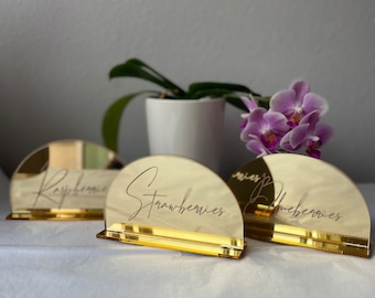 May include: Three gold mirrored table number holders with the words "Raspberries", "Strawberries", and "Blueberries" written in cursive on each.