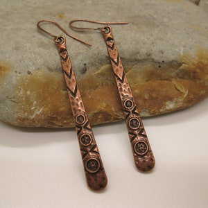 May include: A pair of long, copper earrings with a hammered texture and geometric designs. The earrings have a hook closure.