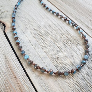 May include: A beaded necklace featuring alternating blue and brown beads. The necklace has a delicate design with small, round beads strung together. The beads are accented with copper-colored spacers, creating a subtle contrast. The necklace is displayed on a wooden surface.
