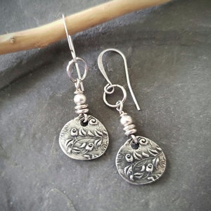 May include: A pair of silver-toned dangle earrings. Each earring features a circular, textured pendant with a floral design. The pendants are connected to a hook with a small bead and coiled wire detail. The earrings are displayed on a dark gray surface.