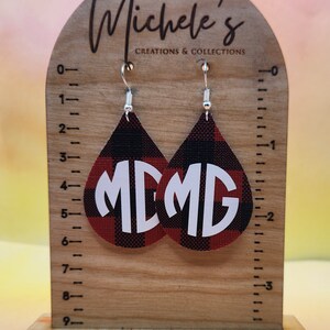 Op de afbeelding: Een paar rode en zwarte buffalo plaid oorbellen met witte monogram initialen "MD". De oorbellen hangen aan zilveren haken aan een houten displaystandaard met een liniaal.