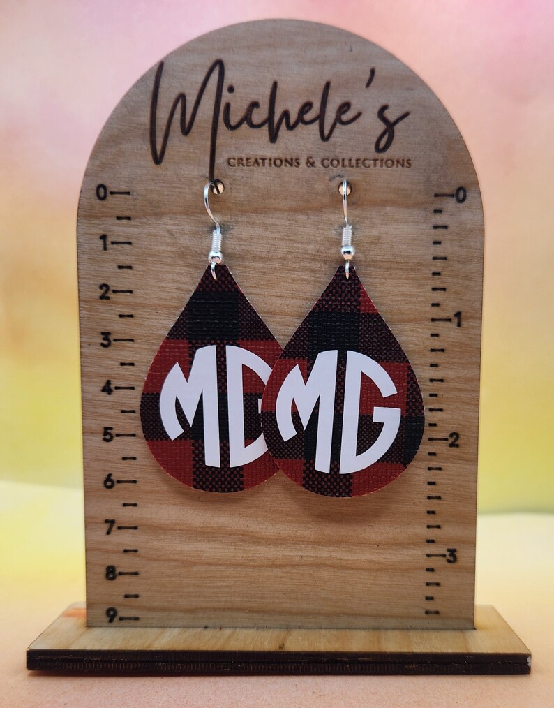 Op de afbeelding: Een paar rode en zwarte buffalo plaid oorbellen met witte monogram initialen "MD". De oorbellen hangen aan zilveren haken aan een houten displaystandaard met een liniaal.