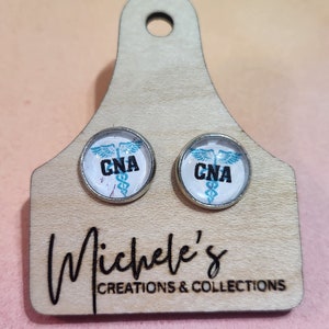 May include: Pair of silver stud earrings with a white background and a blue and black medical caduceus symbol with the letters CNA. The earrings are displayed on a wooden tag with the text "Michele's Creations & Collections".