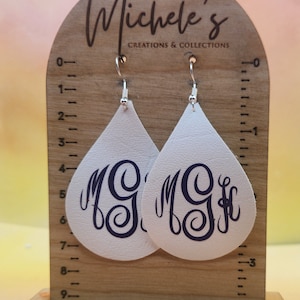 Op de afbeelding: Witte leren oorbellen met een blauw monogram op elke oorbel. De oorbellen worden gepresenteerd op een houten standaard met een liniaal.