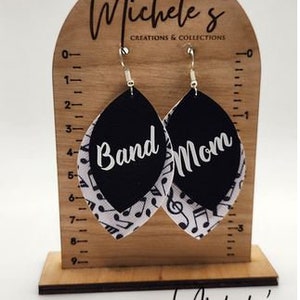 Op de afbeelding: Een paar zwarte en witte oorhangers met de woorden "Band Mom" erop. De oorbellen worden getoond op een houten standaard met een liniaal.