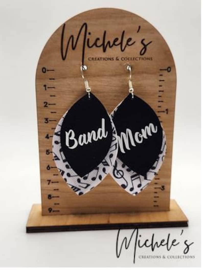 Op de afbeelding: Een paar zwarte en witte oorhangers met de woorden "Band Mom" erop. De oorbellen worden getoond op een houten standaard met een liniaal.