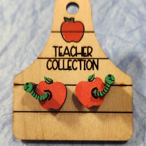 May include: A pair of wooden earrings shaped like red apples with green leaves and a green worm on each apple. The earrings are displayed on a wooden tag that says "Teacher Collection".