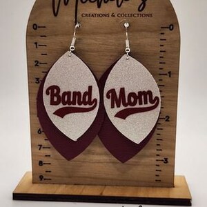 Op de afbeelding: Een paar hangende oorbellen met een witte glitterachtergrond en een donkerrode leren achterkant. De oorbellen hebben de woorden "Band Mom" in zilveren letters.