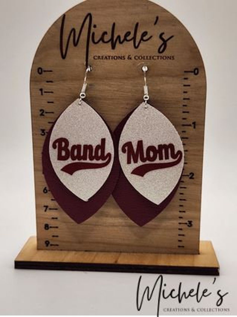 Op de afbeelding: Een paar hangende oorbellen met een witte glitterachtergrond en een donkerrode leren achterkant. De oorbellen hebben de woorden "Band Mom" in zilveren letters.