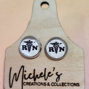 May include: A pair of silver-rimmed stud earrings featuring the R.N. symbol, set against a white background. The earrings are displayed on a wooden tag with the text "Michele's Creations & Collections" etched in black.