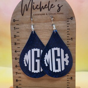 Op de afbeelding: Een paar blauwe glitter-druppelvormige oorbellen met witte monogram-initialen "MG" op elke oorbel. De oorbellen worden getoond op een houten standaard met een liniaal.
