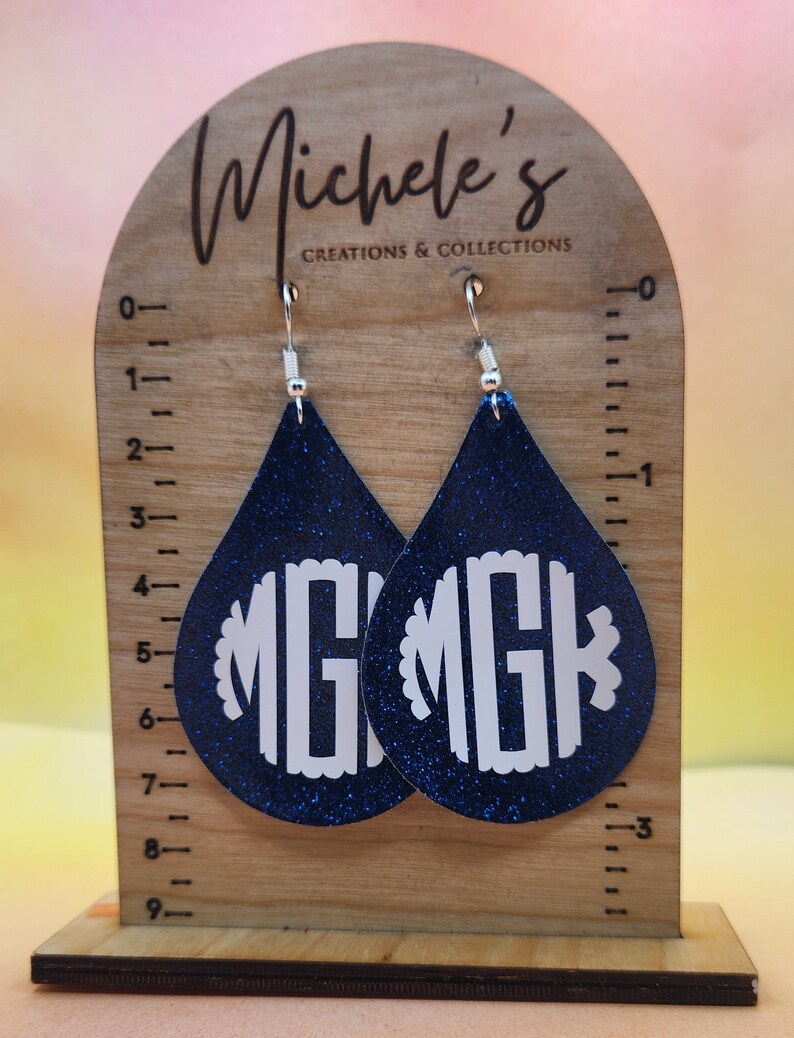 Op de afbeelding: Een paar blauwe glitter-druppelvormige oorbellen met witte monogram-initialen "MG" op elke oorbel. De oorbellen worden getoond op een houten standaard met een liniaal.