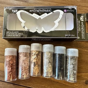 May include: A set of six clear plastic jars filled with metallic mica flakes in copper, rose gold, gold, light gold, black, and silver, accompanied by a black rectangular box labeled Art Ingredients Mica Flakes with a butterfly cutout.