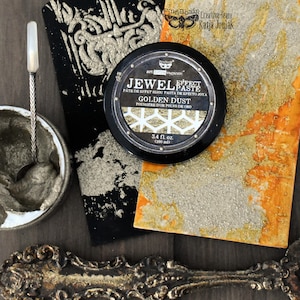May include: A round black container of Art Extravagance Jewel Effect Paste in Golden Dust, 3.4 fl. oz. (100 ml), featuring a white geometric pattern on the label. Beside it are textured black and orange surfaces, a metal spoon, and a decorative object.