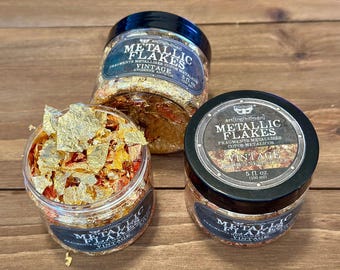 May include: Three clear plastic jars filled with metallic flakes in shades of gold and copper. Each jar features a black lid and a label reading Metallic Flakes Vintage 5 fl. oz. One jar is open, revealing the textured, thin, reflective flakes inside.
