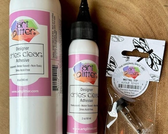 May include: Two white bottles of Art Glitter Designer Dries Clear Adhesive, one 8 oz bottle and one 2 oz bottle, alongside a small clear plastic bag containing a metal precision application tip with a black cap and a dragonfly-themed label.