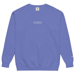 May include: A periwinkle blue crewneck sweatshirt with the text "FILMMAKER IN PROGRESS" in white. The sweatshirt has long sleeves and a comfortable, casual style. The brand label is visible at the top.