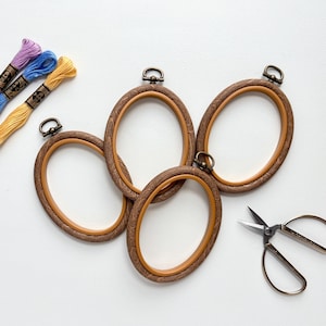 May include: Four oval wooden embroidery hoops with a dark wood grain finish and brass hardware are arranged on a white surface. Three spools of colorful embroidery floss and a pair of scissors are also present, suggesting a crafting theme.