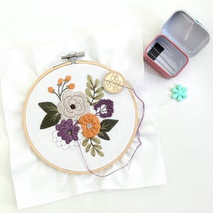 K&ouml;nnte beinhalten: Nahaufnahme eines floralen Stickrahmens mit wei&szlig;em Hintergrund und bunten Blumen. Der Rahmen ist aus Holz und hat einen Metallring am Rand. Die Blumen sind in Violett-, Orange- und Gr&uuml;nt&ouml;nen gestickt.