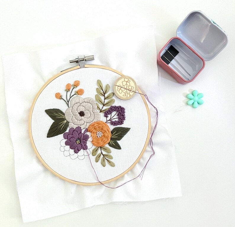 K&ouml;nnte beinhalten: Nahaufnahme eines floralen Stickrahmens mit wei&szlig;em Hintergrund und bunten Blumen. Der Rahmen ist aus Holz und hat einen Metallring am Rand. Die Blumen sind in Violett-, Orange- und Gr&uuml;nt&ouml;nen gestickt.