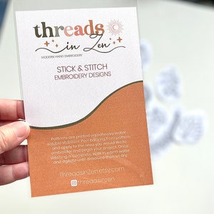 Puede incluir: Una funda de pl&aacute;stico transparente que contiene una tarjeta con el texto "threads in Zen" en marr&oacute;n y rosa. La tarjeta dice "STICK & STITCH EMBROIDERY DESIGNS" e incluye instrucciones. El fondo muestra dise&ntilde;os de bordado blancos.