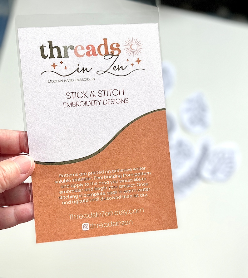 Puede incluir: Una funda de pl&aacute;stico transparente que contiene una tarjeta con el texto "threads in Zen" en marr&oacute;n y rosa. La tarjeta dice "STICK & STITCH EMBROIDERY DESIGNS" e incluye instrucciones. El fondo muestra dise&ntilde;os de bordado blancos.