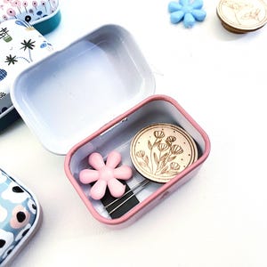 K&ouml;nnte beinhalten: Eine kleine rosafarbene Metalldose mit einem Blumendesign auf dem Deckel. Die Dose ist ge&ouml;ffnet und enth&auml;lt einen rosafarbenen Blumenknopf, zwei N&auml;hnadeln und einen runden Holzknopf mit einem Blumendesign.