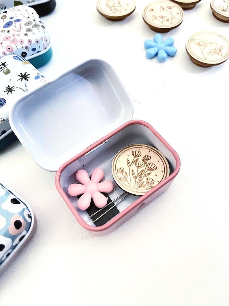 K&ouml;nnte beinhalten: Eine kleine rosafarbene Metalldose mit einem Blumendesign auf dem Deckel. Die Dose ist ge&ouml;ffnet und enth&auml;lt einen rosafarbenen Blumenknopf, zwei N&auml;hnadeln und einen runden Holzknopf mit einem Blumendesign.