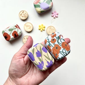 May include: Four small, rectangular, metal tins with floral designs. The tins have different floral patterns, including purple flowers, orange flowers, and green leaves. The tins are perfect for storing small items like jewelry, buttons, or sewing supplies.