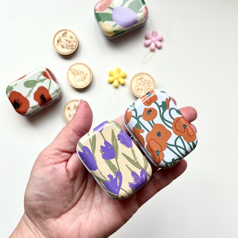 May include: Four small, rectangular, metal tins with floral designs. The tins have different floral patterns, including purple flowers, orange flowers, and green leaves. The tins are perfect for storing small items like jewelry, buttons, or sewing supplies.