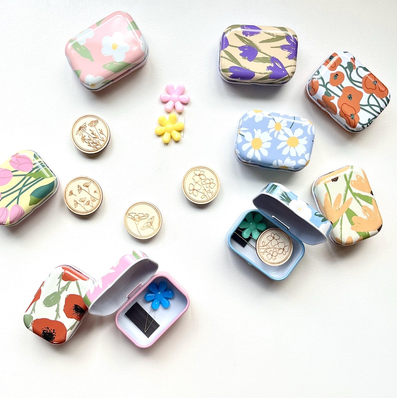 May include: Assortment of small, rectangular, floral patterned metal tins with lids. Some tins are open and contain small wooden stamps with floral designs.  A few colorful plastic flower embellishments are scattered around the tins.