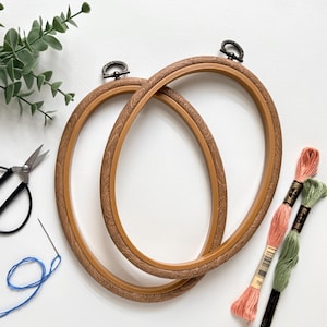 May include: Two oval embroidery hoops with a wood grain pattern. The hoops are brown with a metal clasp. Also pictured are embroidery floss in shades of peach and green, a needle with blue thread, and black scissors.