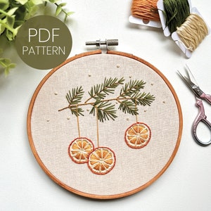 May include: Embroidered hoop art featuring a green pine branch with three orange slices hanging from it. The background is a cream-colored fabric with white dots. The hoop is made of wood and has a natural finish.