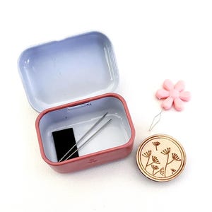 K&ouml;nnte beinhalten: Ein rosafarbenes N&auml;hset aus Metall mit einer kleinen schwarzen Garnrolle, zwei Nadeln und einem Holz-Knopf mit Blumenmuster. Das Set enth&auml;lt auch einen rosafarbenen Plastik-Nadelfadenzieher mit Blumenmuster.