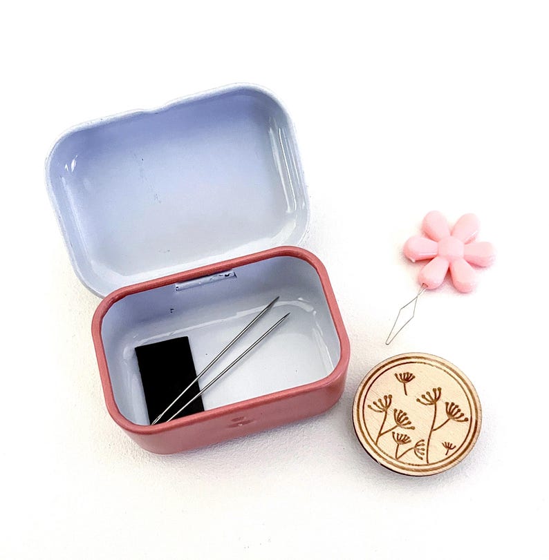 K&ouml;nnte beinhalten: Ein rosafarbenes N&auml;hset aus Metall mit einer kleinen schwarzen Garnrolle, zwei Nadeln und einem Holz-Knopf mit Blumenmuster. Das Set enth&auml;lt auch einen rosafarbenen Plastik-Nadelfadenzieher mit Blumenmuster.