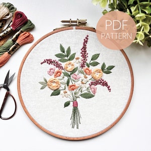 May include: Embroidered floral bouquet in a hoop with a beige background. The bouquet features pink, orange, and yellow flowers with green leaves and stems. The hoop is made of wood and has a brown finish. The text "PDF PATTERN" is visible in the upper right corner of the image.