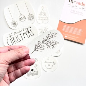Puede incluir: Una colecci&oacute;n de dise&ntilde;os de bordado de pegar y coser blancos con arte lineal negro. Los dise&ntilde;os incluyen adornos navide&ntilde;os, un &aacute;rbol de Navidad, las palabras "Merry Christmas", una ramita de pino, una ramita de acebo, un oso y una taza de caf&eacute;.