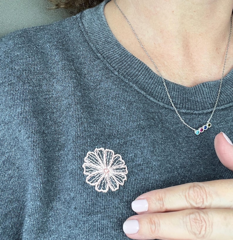 Puede incluir: Un broche de flores calado rosa claro prendido a una sudadera gris. Tambi&eacute;n se ve un delicado collar de plata con tres peque&ntilde;as piedras preciosas de colores. El broche es de dise&ntilde;o floral, y el collar es de estilo sencillo y elegante.