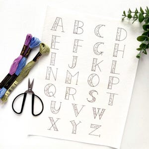 May include: A sheet of paper with the alphabet, each letter designed with decorative flourishes. Embroidery floss in purple, blue, and green, along with small black scissors, are arranged to the left. A sprig of greenery is in the upper right corner.